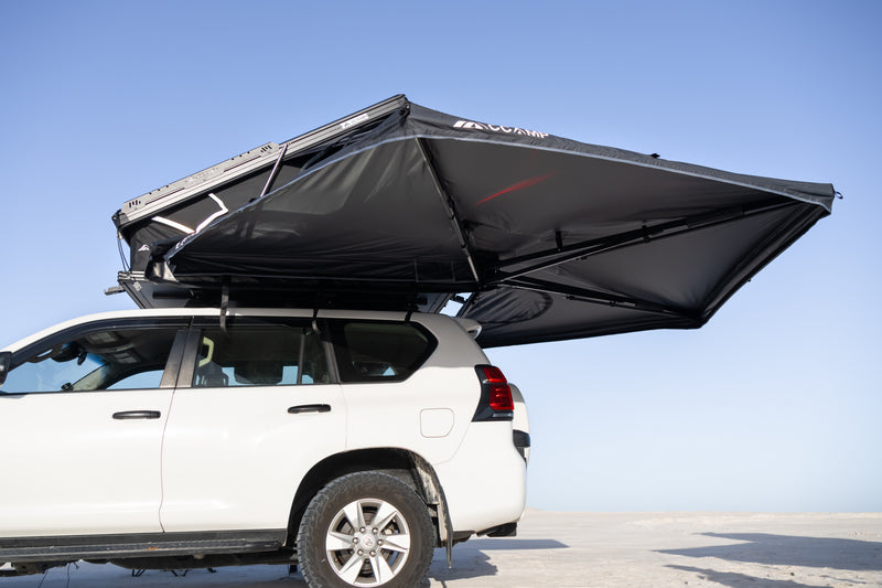 Load image into Gallery viewer, A white SUV parked outdoors with a large black rooftop tent canopy extended over the roof, shown against a clear blue sky.
