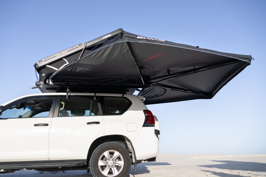 A white SUV parked outdoors with a large black rooftop tent canopy extended over the roof, shown against a clear blue sky.