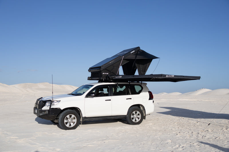 Load image into Gallery viewer, A white SUV parked in a desert landscape with sand dunes under a clear blue sky, equipped with a black rooftop tent mounted on the roof.
