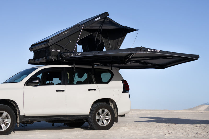 Load image into Gallery viewer, A white SUV parked outdoors on a desert surface with a black rooftop tent and matching awning extended over the roof against a clear blue sky.

