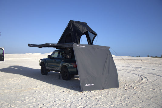 A dark green SUV parked on white sand dunes with a black rooftop tent and gray ground canopy tent attached, under a clear blue sky.
