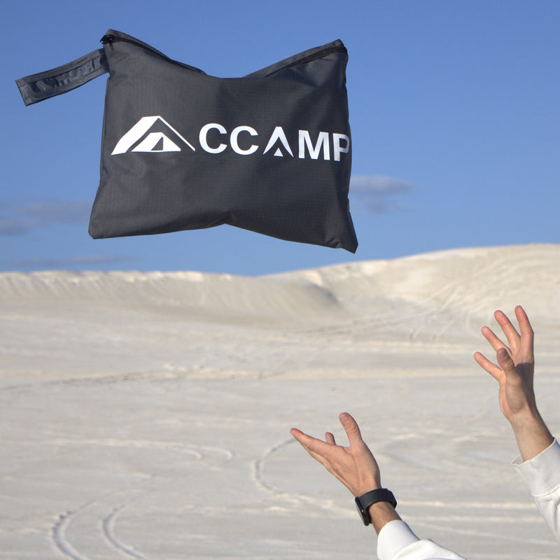 Load image into Gallery viewer, A black CCAMP sandbag with a white logo is shown mid-air being thrown by a person whose hands and arms are visible against a background of white sand dunes and a clear blue sky.
