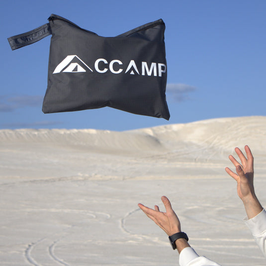 A black CCAMP sandbag with a white logo is shown mid-air being thrown by a person whose hands and arms are visible against a background of white sand dunes and a clear blue sky.