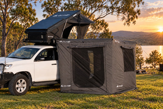 A black CCAMP rooftop tent mounted on a white pickup truck, parked outdoors on grass near a lake at sunset with trees and hills in the background.