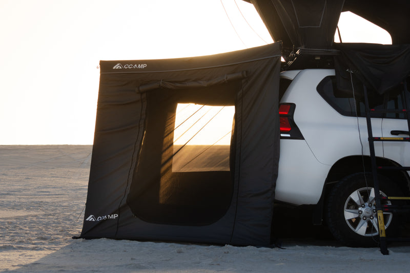 Load image into Gallery viewer, A black CCAMP car awning tent attached to the side of a white SUV on a flat desert landscape at sunset.
