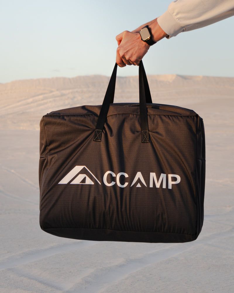 Load image into Gallery viewer, A person holds up a large black CCAMP carrying bag with white logo branding, set against a blurred desert landscape background.
