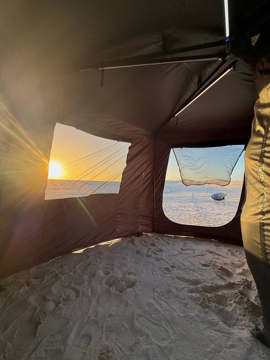 A view from inside a dark brown ice fishing tent on snow, looking out through windows at a snowy outdoor landscape during a bright winter sunset.