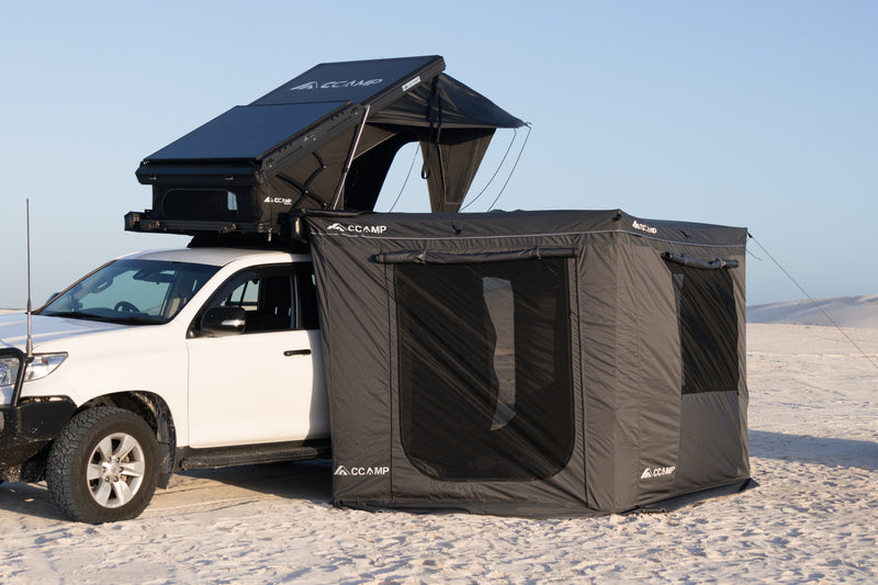 Load image into Gallery viewer, A black CCamp rooftop tent and matching ground tent system attached to a white pickup truck in a sandy desert setting under a clear blue sky.
