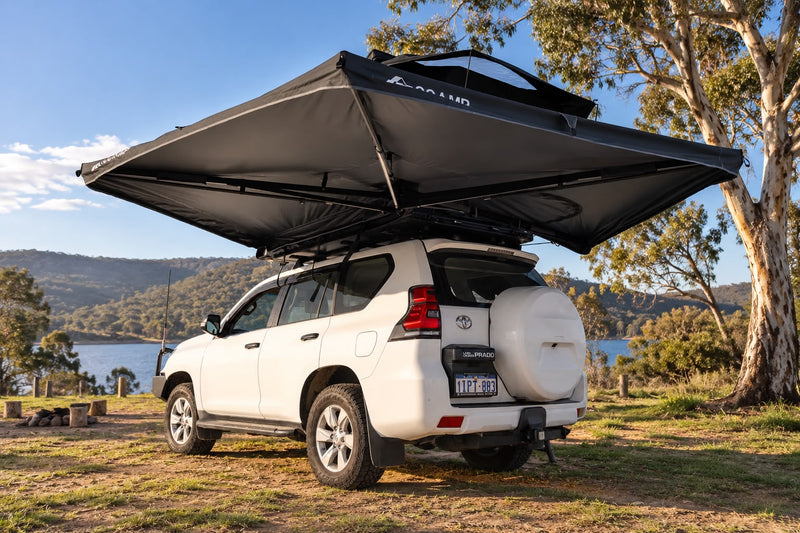 Load image into Gallery viewer, A white SUV parked outdoors by a lake with a large black rooftop canopy tent extended over the roof, showing the rear and side of the vehicle and surrounding trees and hills.
