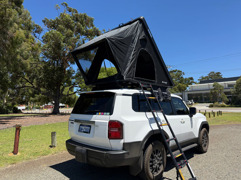 Load image into Gallery viewer, White SUV with a black rooftop tent parked on a road with trees and buildings in the background.
