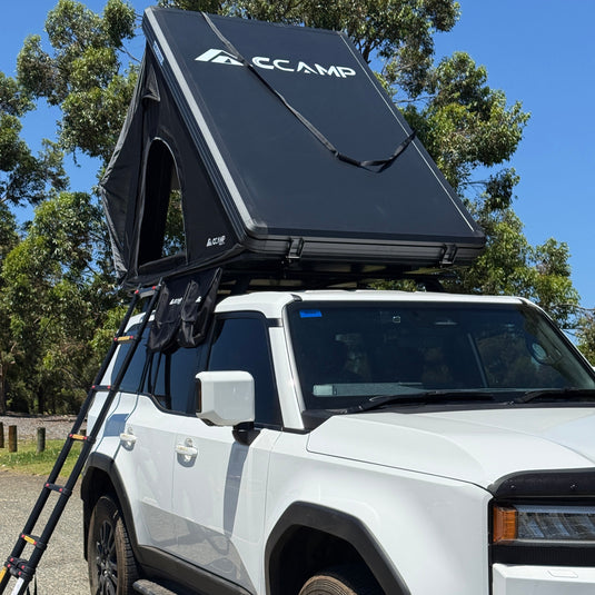 White SUV with a black pop-up camper attached, parked outdoors with trees in the background.