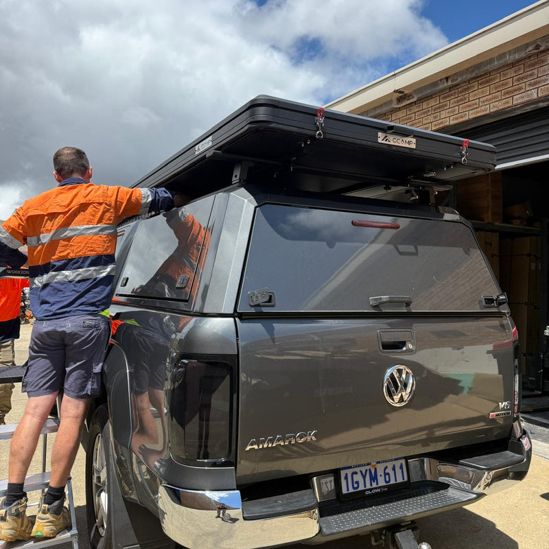 Load image into Gallery viewer, A man in an orange and navy high-visibility work shirt stands beside a dark gray Volkswagen Allroad pickup truck parked outdoors. The truck is fitted with a black roof top box and a dark canopy cover.
