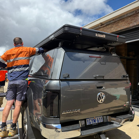 A man in an orange and navy high-visibility work shirt stands beside a dark gray Volkswagen Allroad pickup truck parked outdoors. The truck is fitted with a black roof top box and a dark canopy cover.