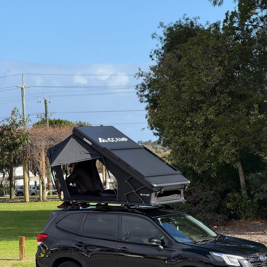 A black rooftop tent mounted on the roof of a black SUV, parked outdoors on a paved area with trees, grass, and a blue sky in the background.